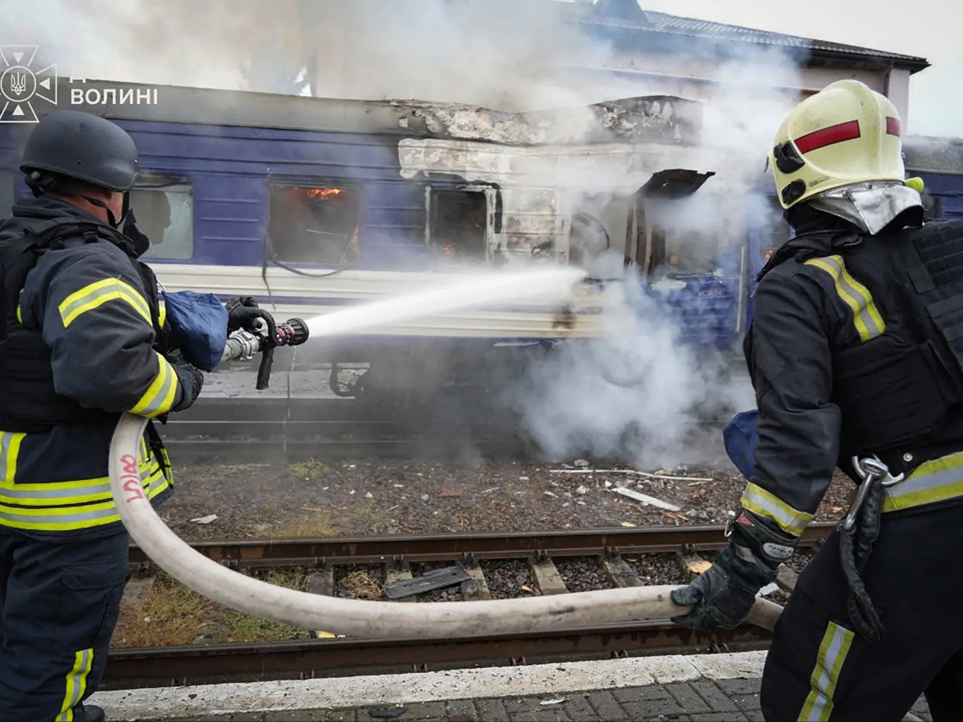 Firefighters spray water on a smoke-filled burning passenger train beside railway tracks.