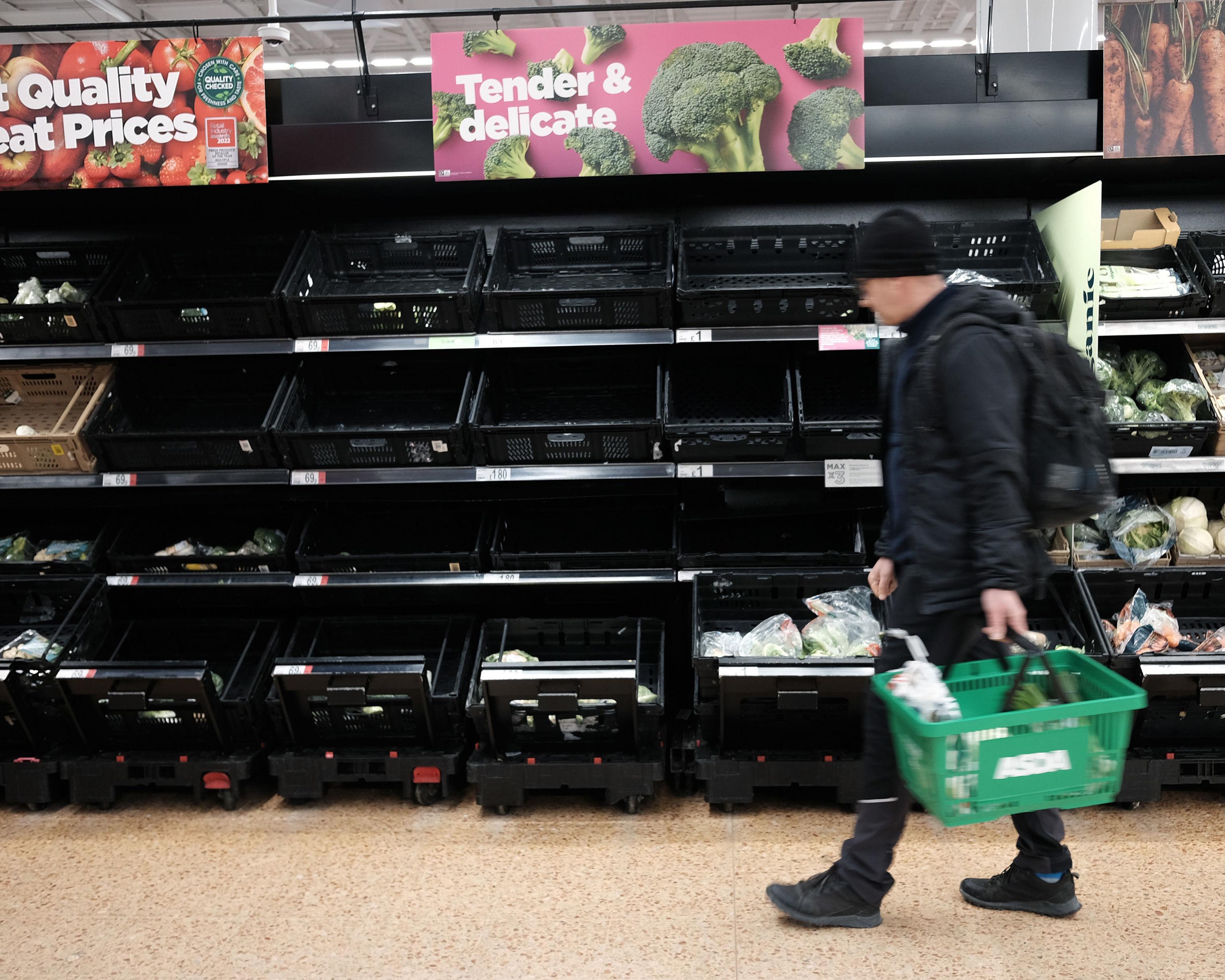 Shopper carrying a green basket passes largely empty produce shelves in a supermarket.