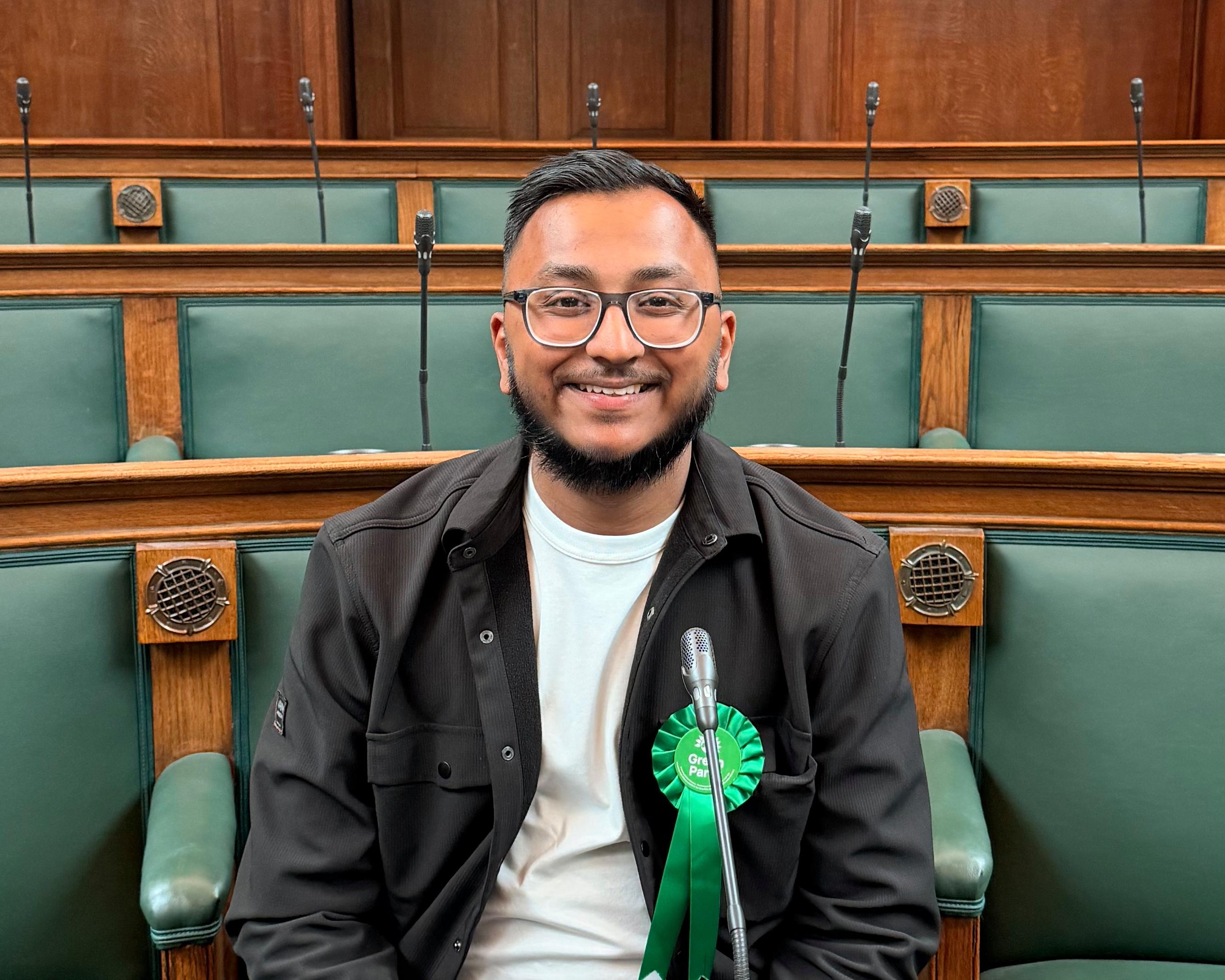 Smiling man with glasses and beard sits at a parliamentary bench beside a microphone.