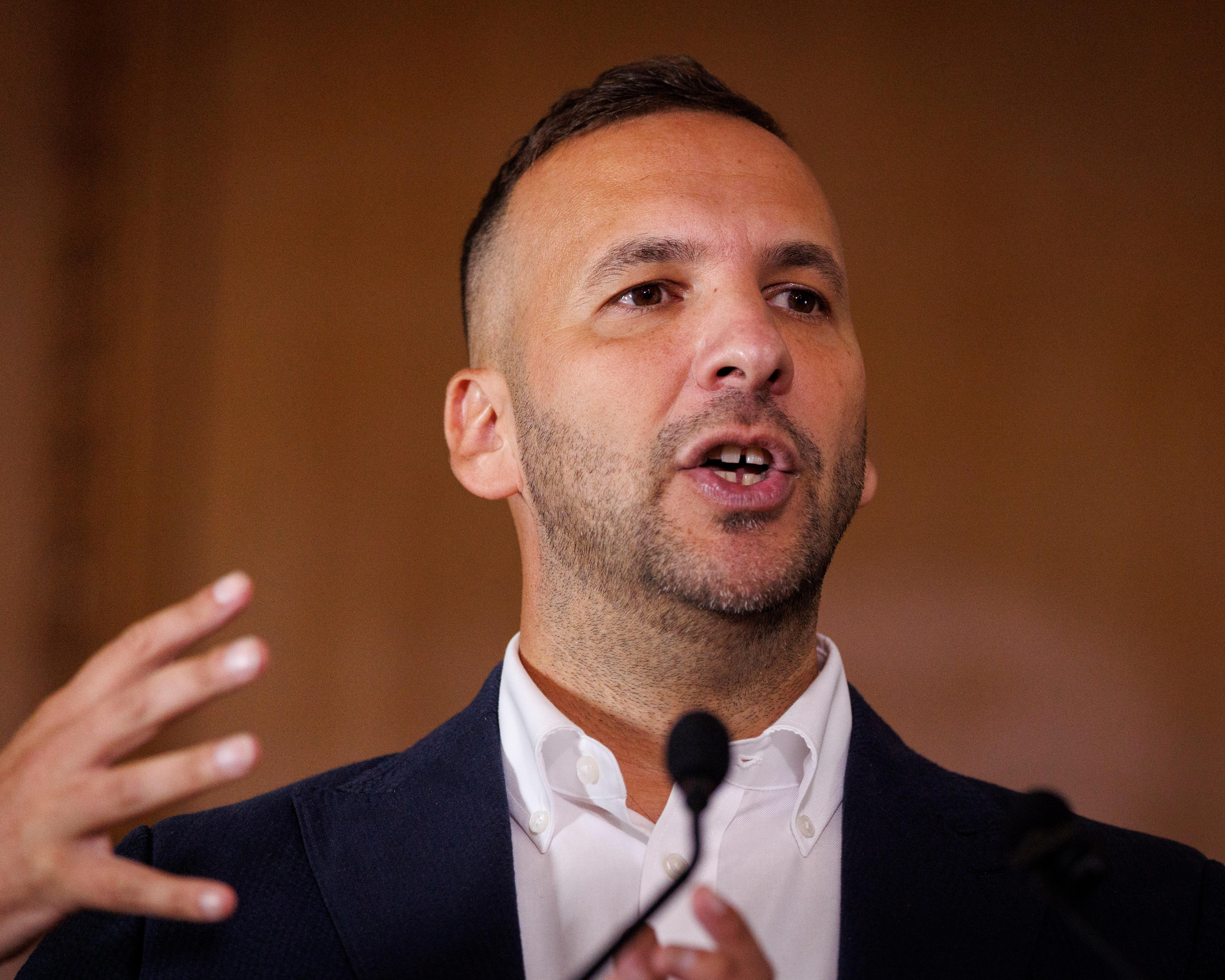 A bearded man in a white shirt and dark jacket speaks into a microphone, gesturing with one hand.