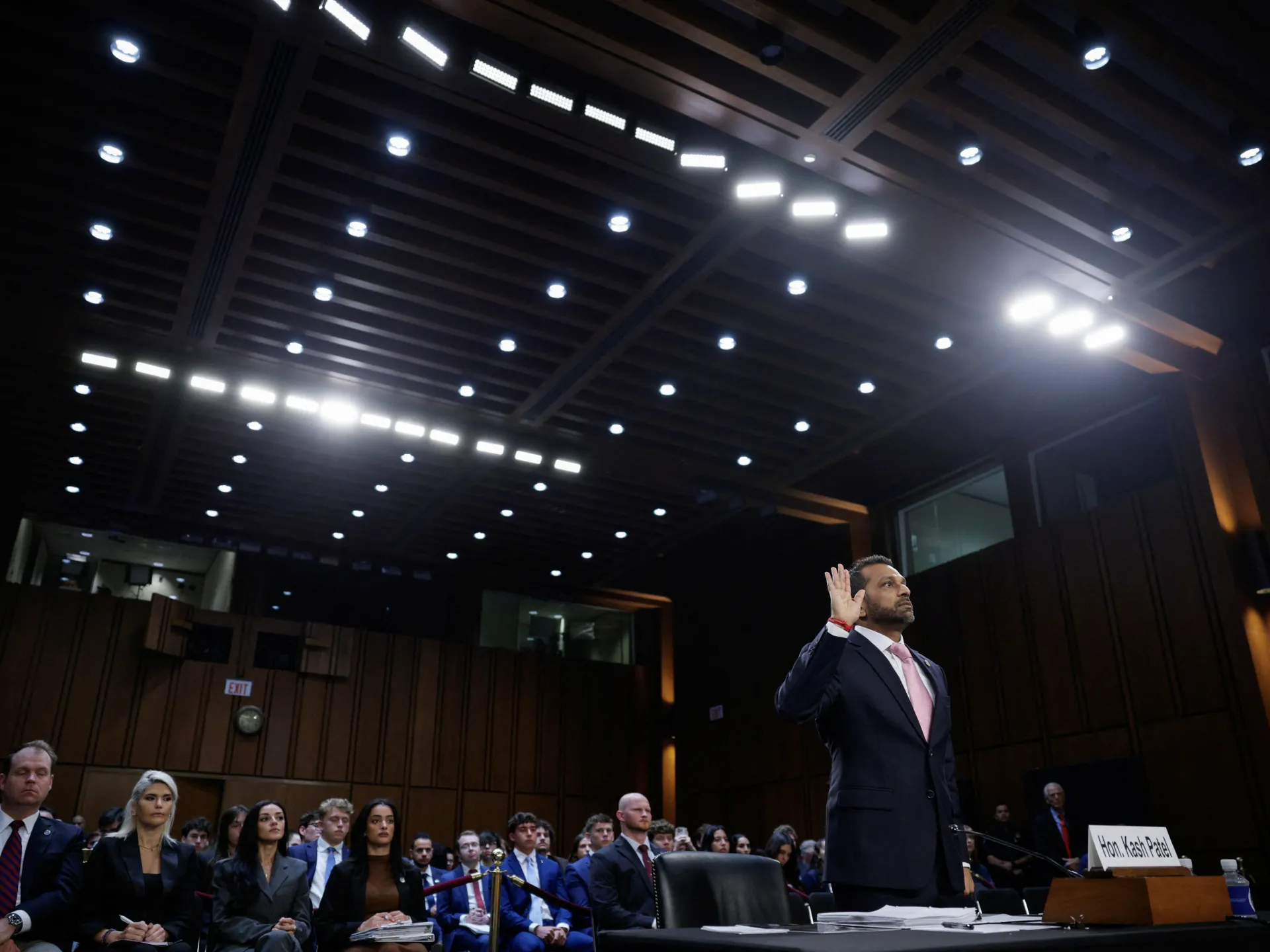 Man in suit raises his hand while testifying before a crowded hearing room.
