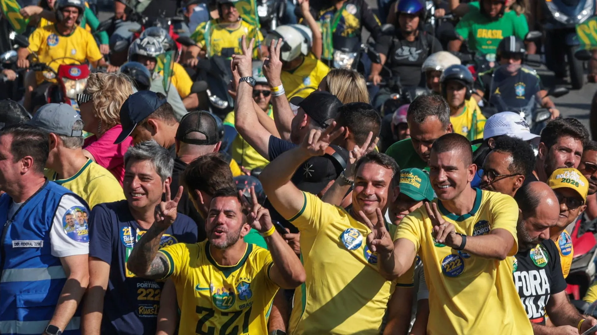 Crowd of people in yellow and green shirts, many wearing helmets, waving and raising peace signs.