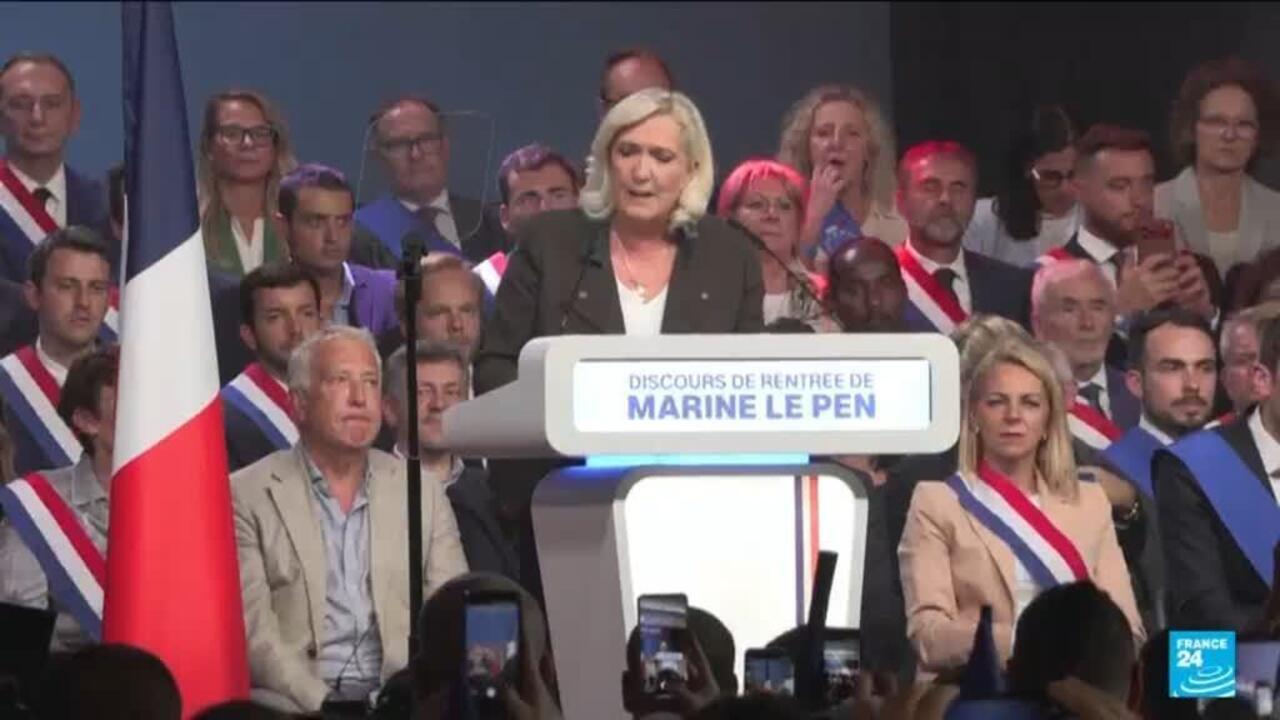 Marine Le Pen speaks at a podium surrounded by French flags and sash-wearing supporters.