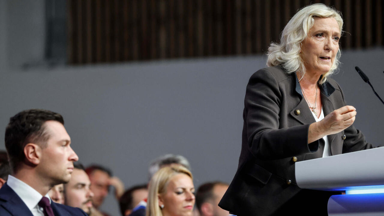 A white-haired woman speaks at a lectern while seated attendees listen in the foreground.
