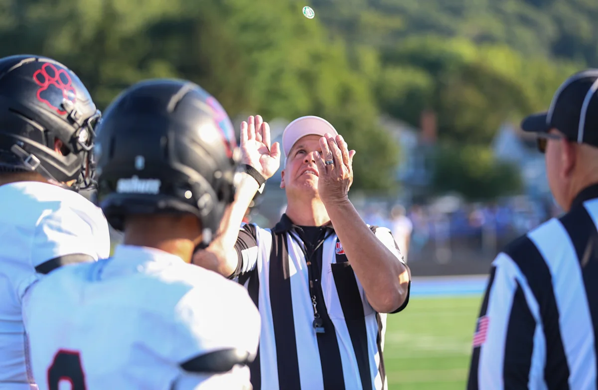 Football official tosses a ball as helmeted players gather during a high school game.