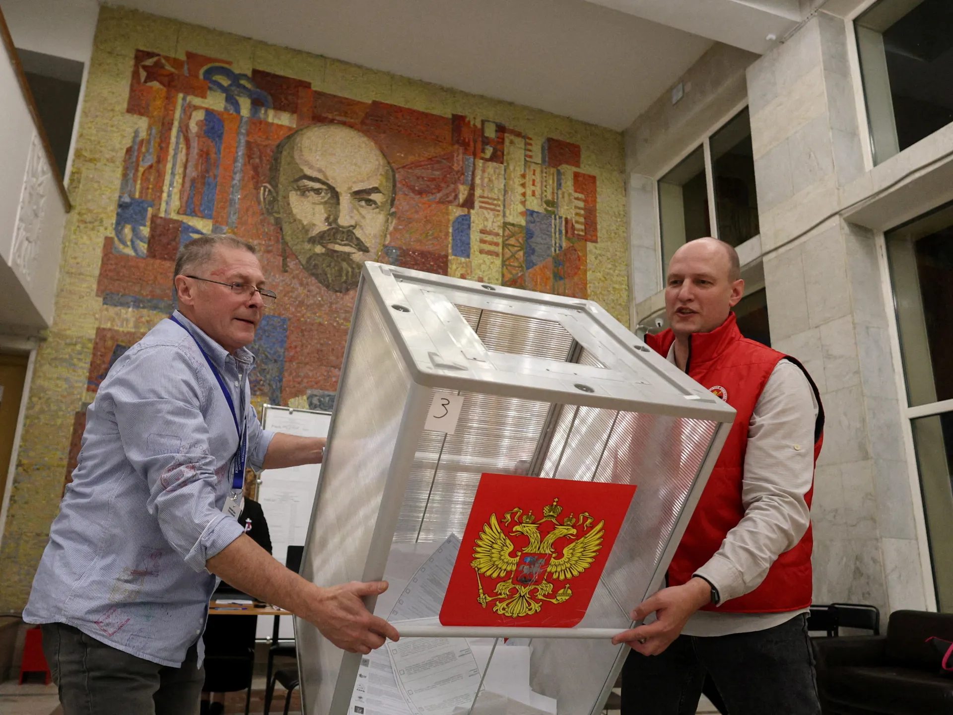 Two men carry a transparent ballot box displaying Russia’s coat of arms beneath a Lenin mosaic.