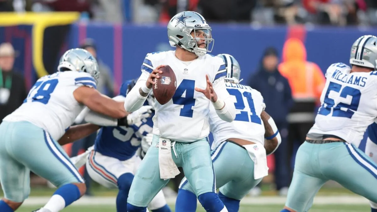 Dallas Cowboys quarterback in helmet holds the football behind his offensive line during a game.
