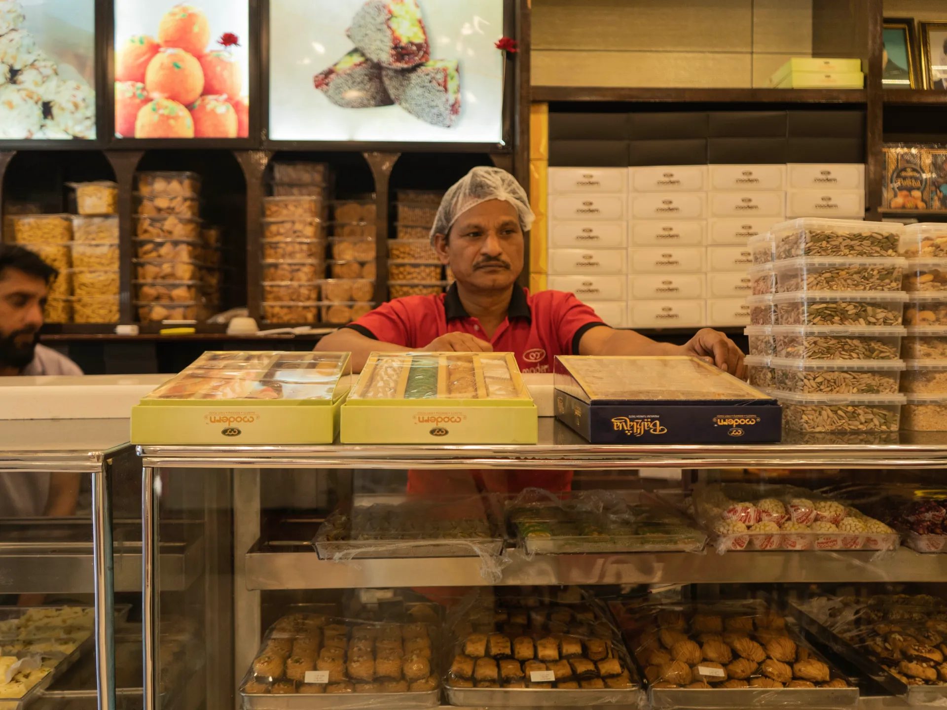 Sweets shop worker stands behind trays of boxed confections, nuts and pastries.