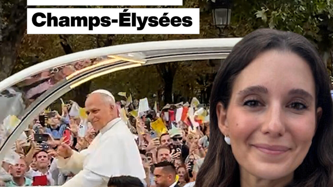 A woman appears beside Pope Leo in a popemobile, with a crowd waving flags and filming.