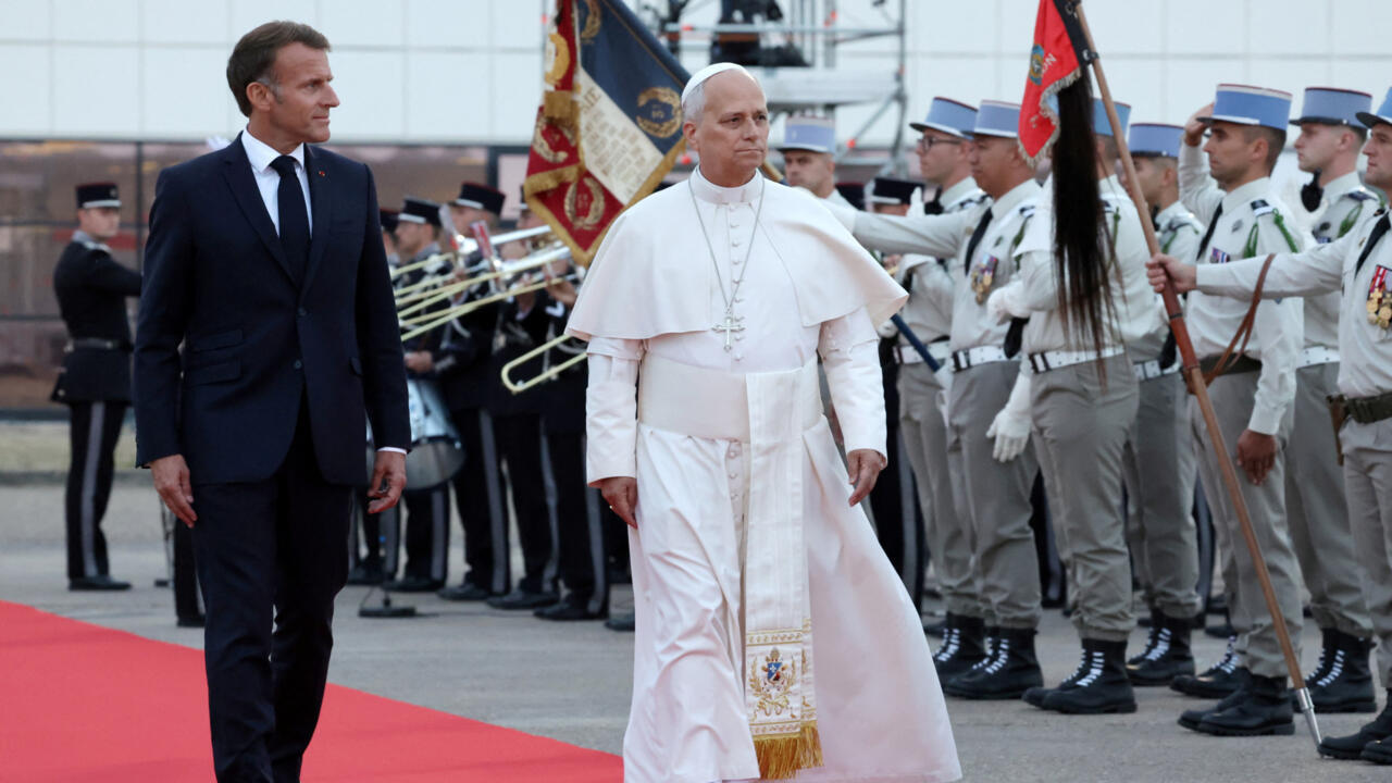 A white-robed cleric walks beside a suited man as uniformed guards stand at attention.
