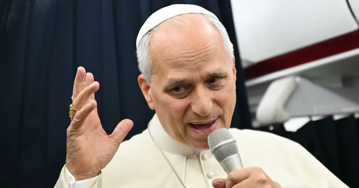A man in white clerical clothing and skullcap speaks into a microphone, raising one hand.