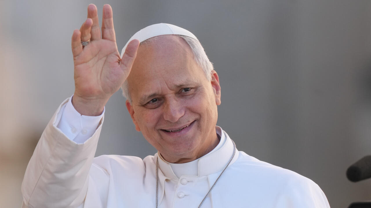 Pope Leo, wearing white papal vestments and skullcap, raises his hand in greeting.
