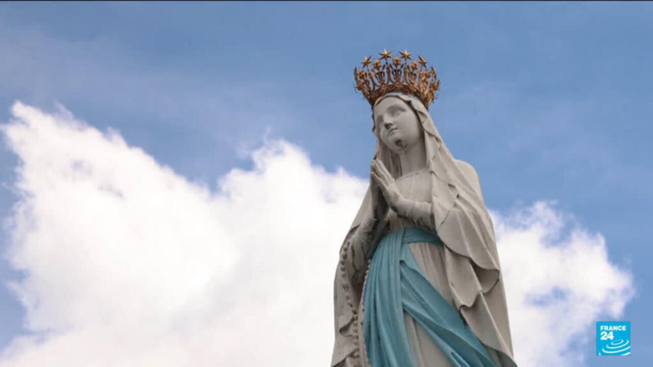 A crowned statue of the Virgin Mary in a blue robe stands against a cloudy sky.