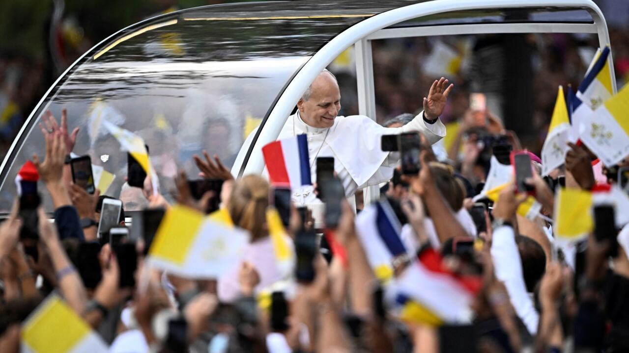A white-robed cleric waves from a vehicle as a crowd raises phones and yellow-and-white flags.