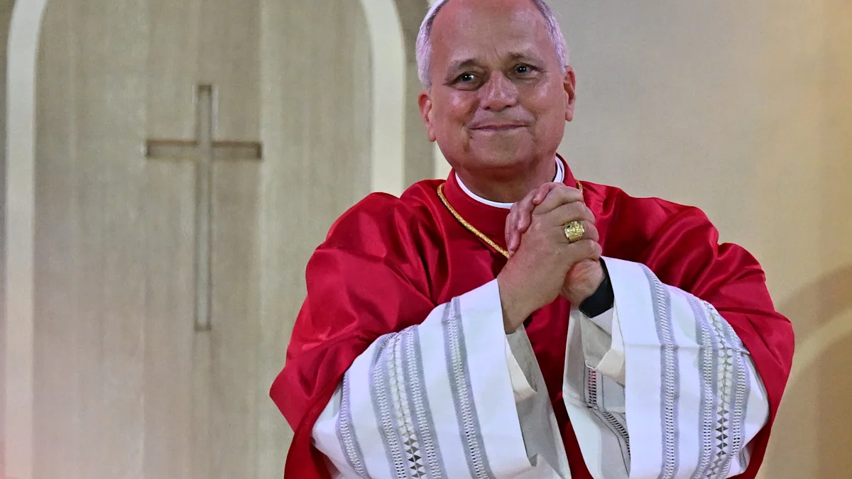 A man in red and white clerical vestments clasps his hands in front of a cross.