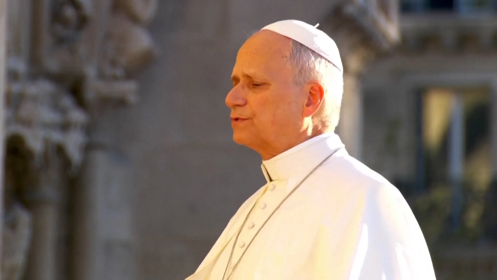 A man in white papal robes and skullcap stands in profile against a blurred background.