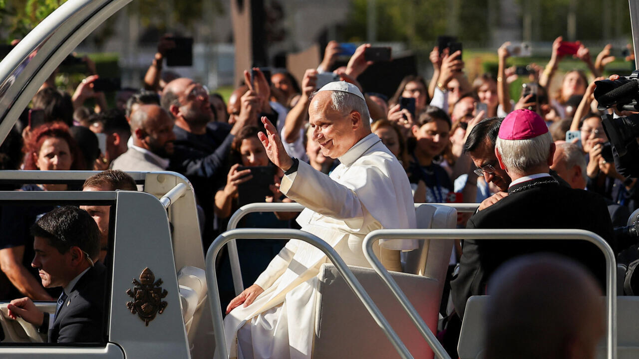 A white-robed, white-capped man rides in a vehicle as a crowd raises phones around him.