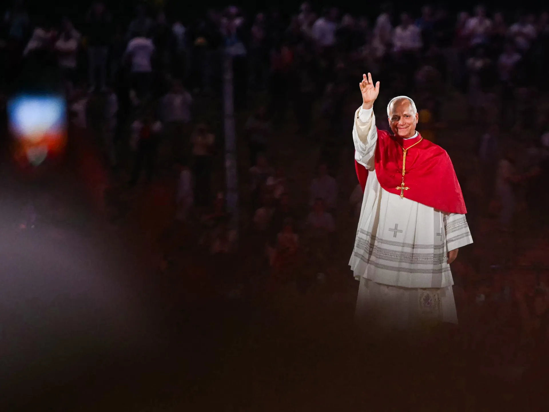 A man in white clerical robes and a red cape waves before a dimly lit crowd.