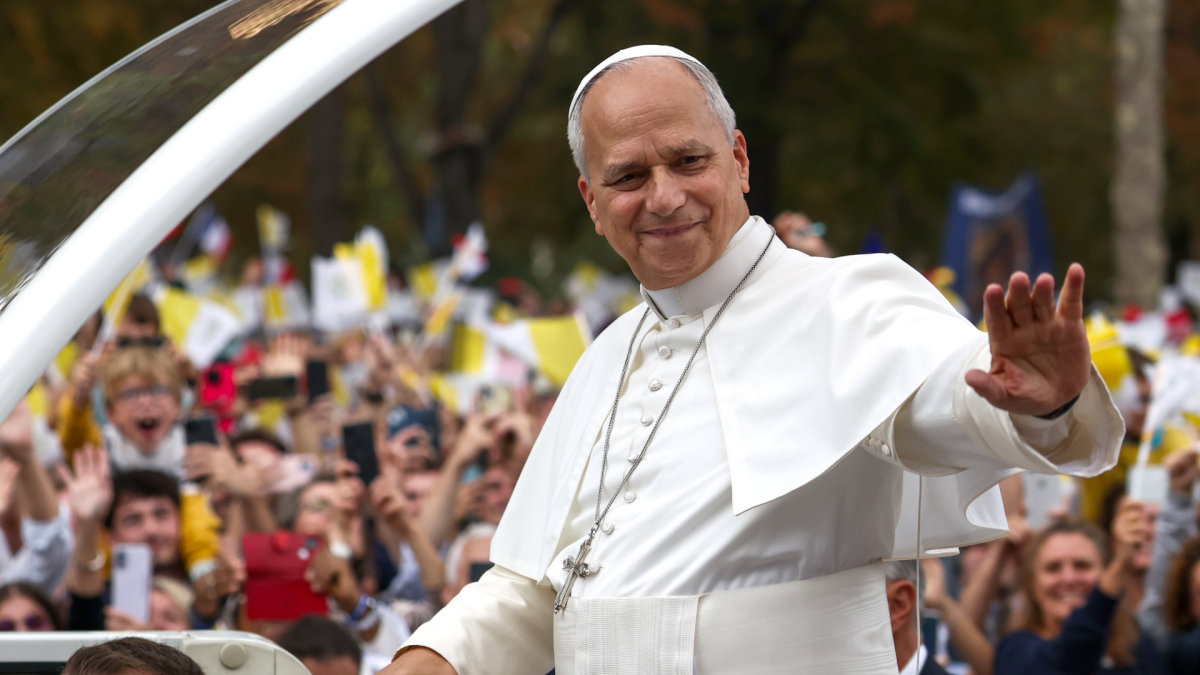 A man in white papal clothing waves from a vehicle as a crowd holds flags and phones nearby.