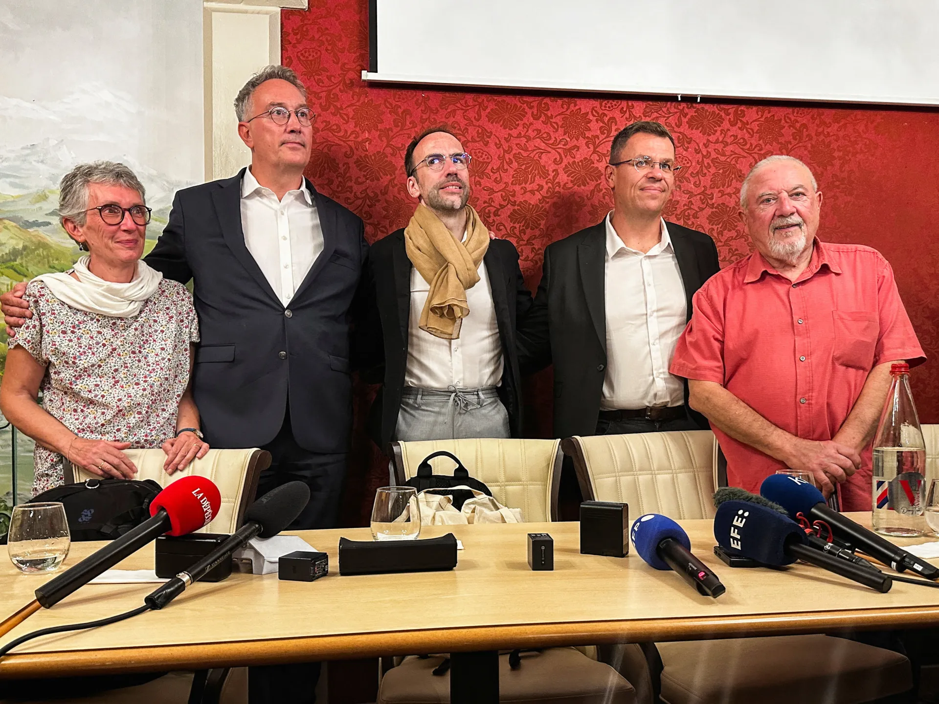 Five adults stand behind a conference table with microphones, bags and water bottles in front of them.