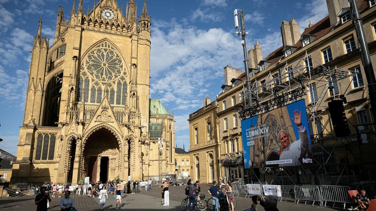 Gothic cathedral and nearby buildings overlook a plaza with crowds, barriers, scaffolding, and a papal event poster.