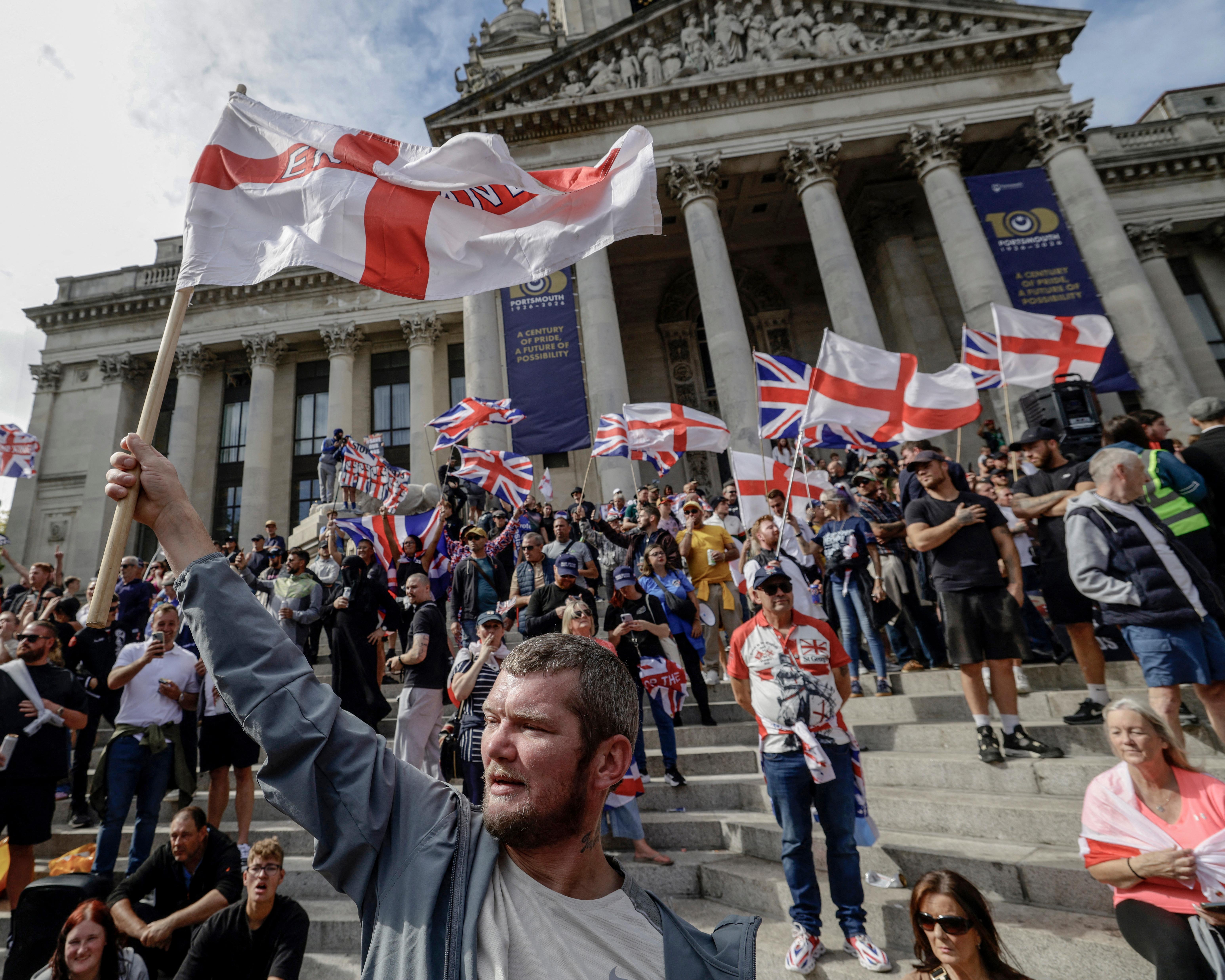 Crowd waving English flags on the steps of a grand neoclassical building.