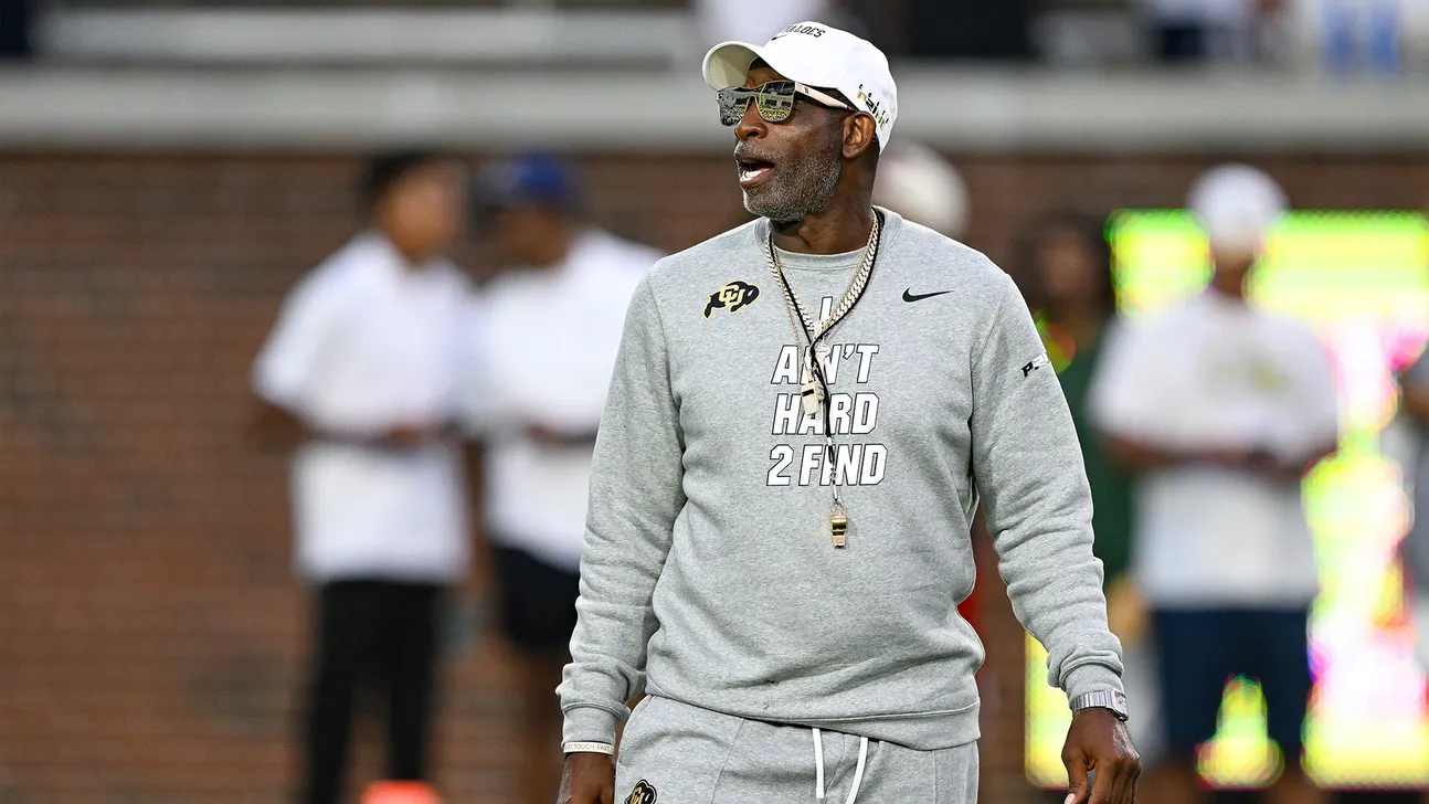 Coach in gray sweatshirt, white cap and sunglasses walking on a football field