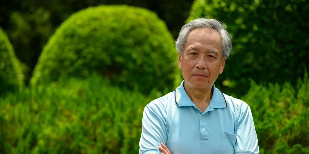 Older man in light blue polo shirt standing outdoors in a lush green garden