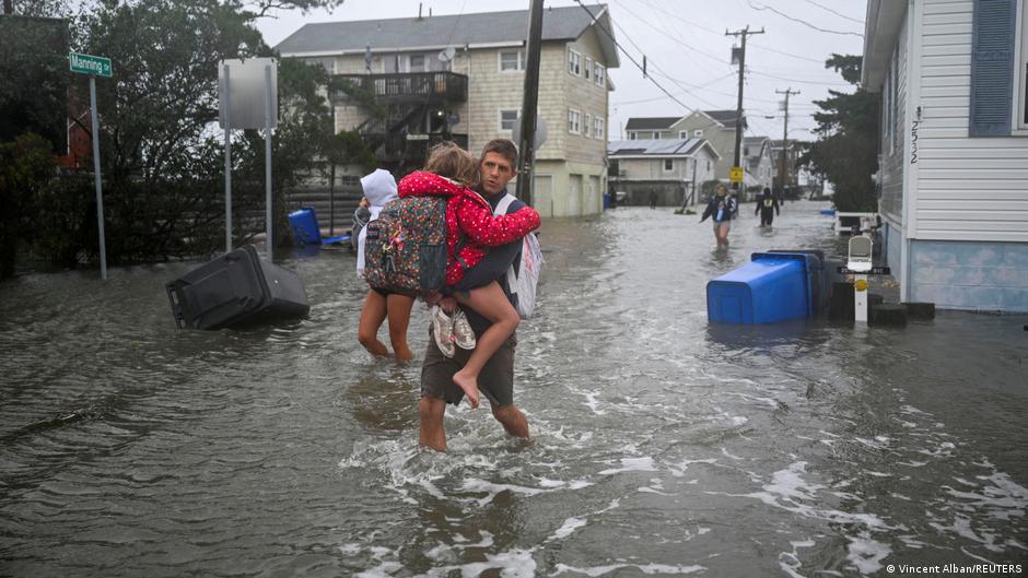 A man carries a child through a flooded residential street as others wade through water nearby.