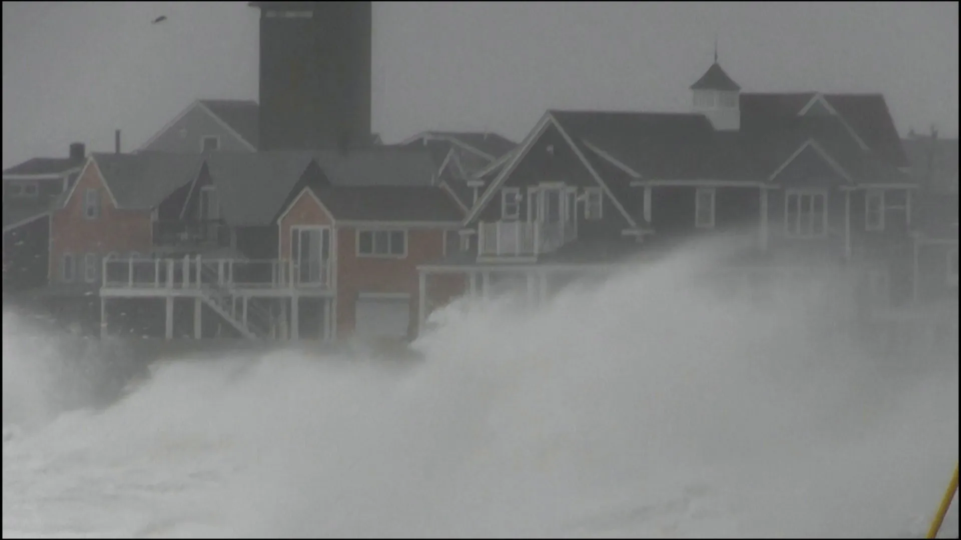 Large waves and spray crash against coastal houses beneath a gray, stormy sky.