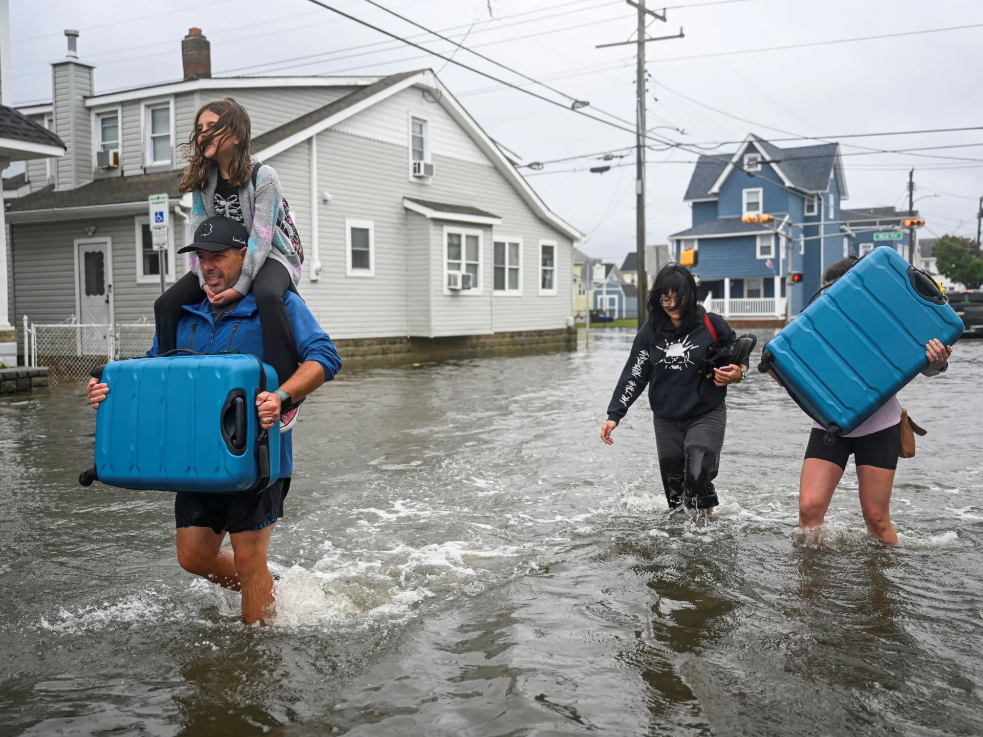 People wade through flooded residential street carrying suitcases, with a child riding on a man's shoulders.