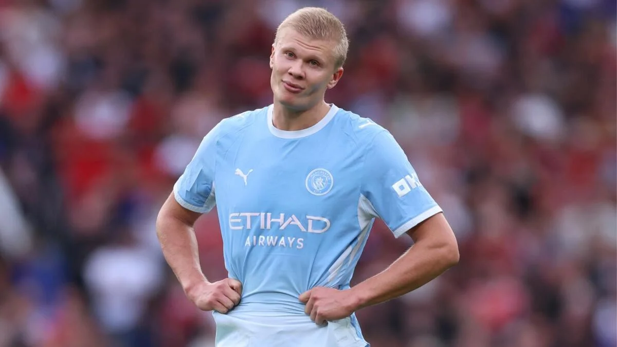 Manchester City player in a sky-blue kit adjusts his shirt against a blurred crowd