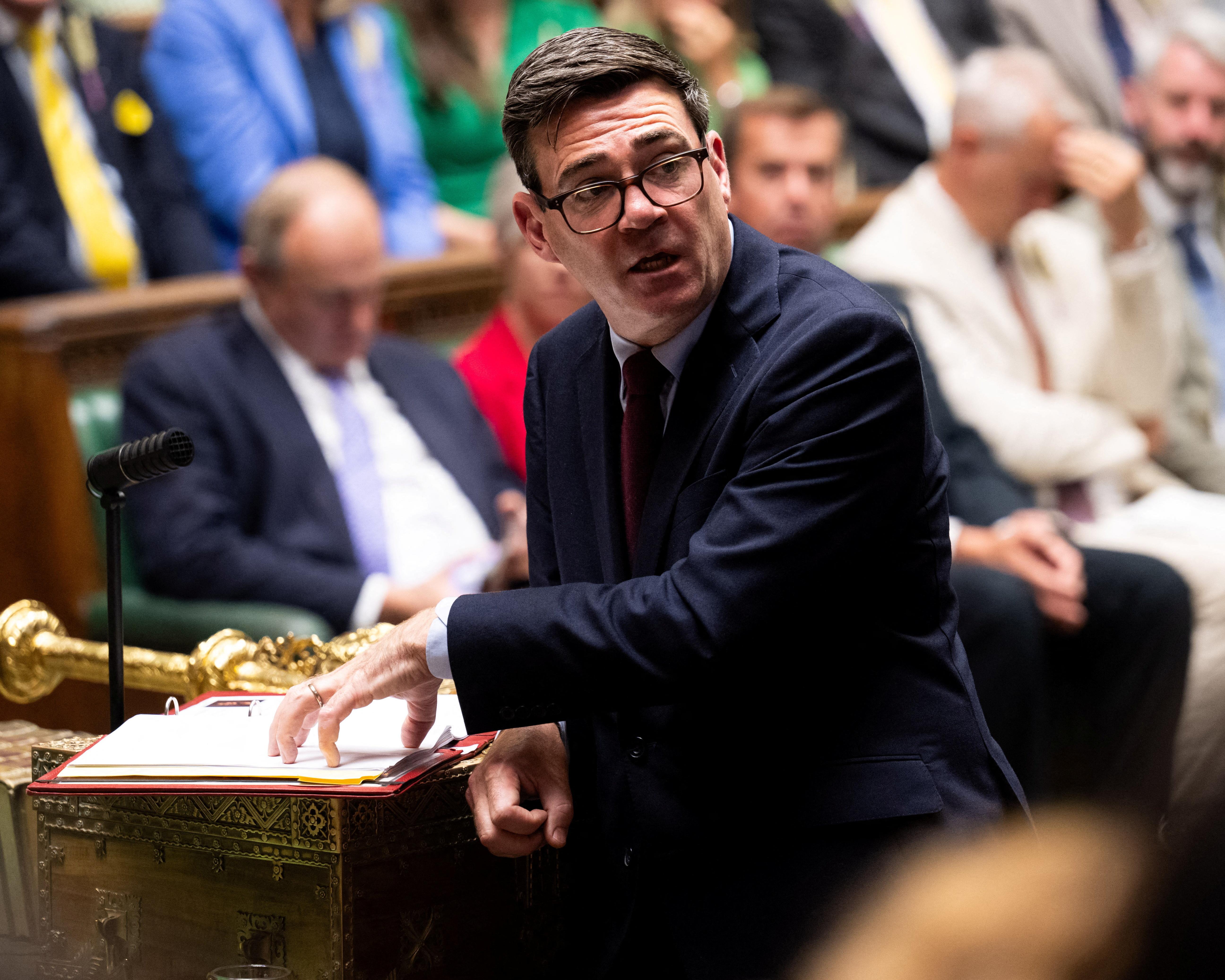 Suited politician speaking at a chamber desk beside papers and a microphone