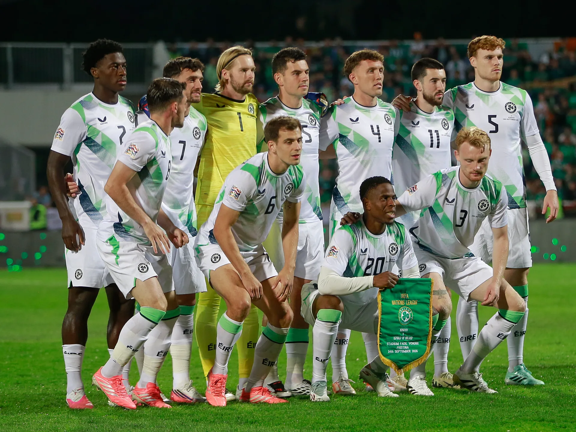 Irish football team players pose together on the pitch, with one holding a green UEFA Nations League pennant.