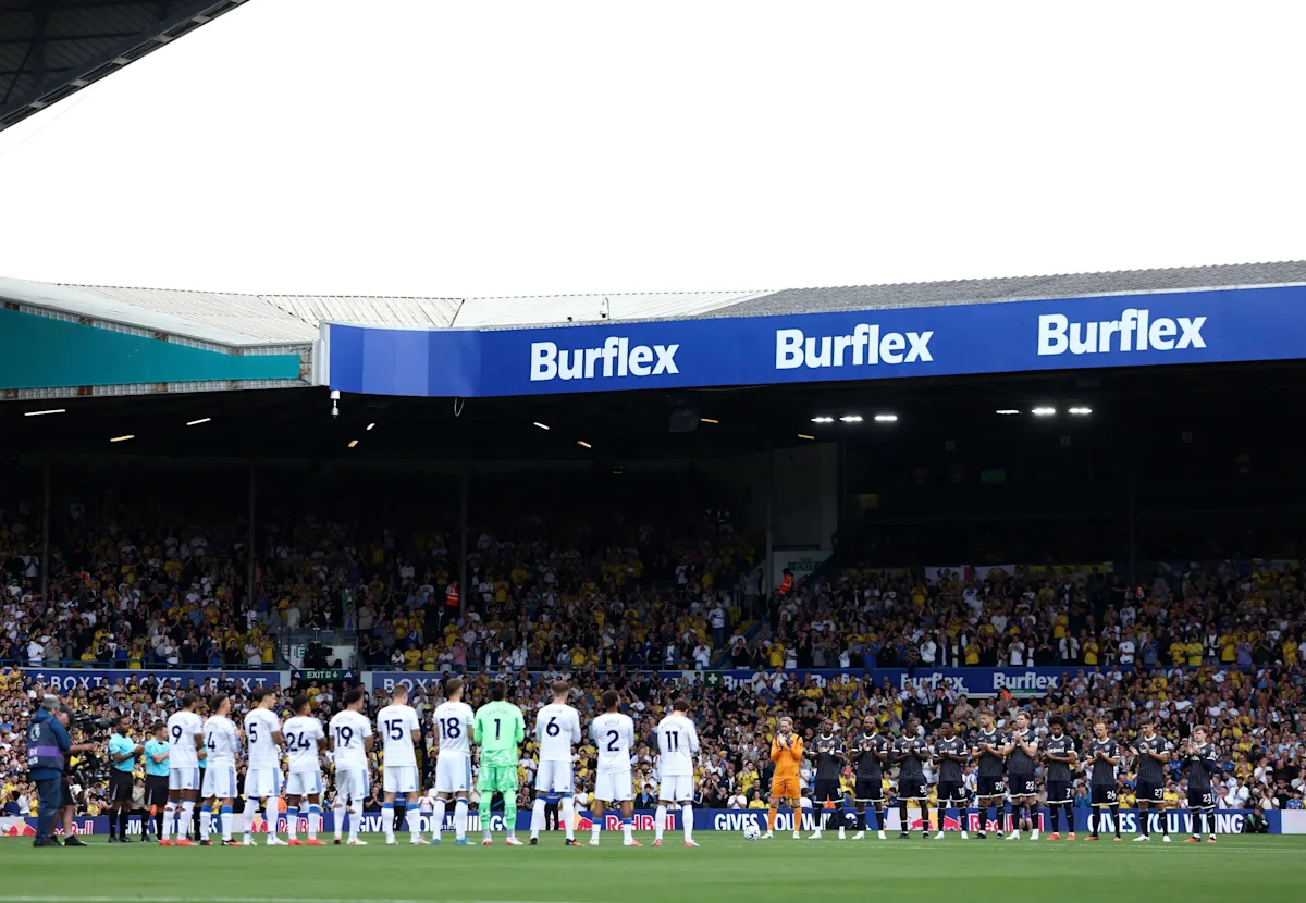 Football teams line up on the pitch before a match in front of a crowded stand.