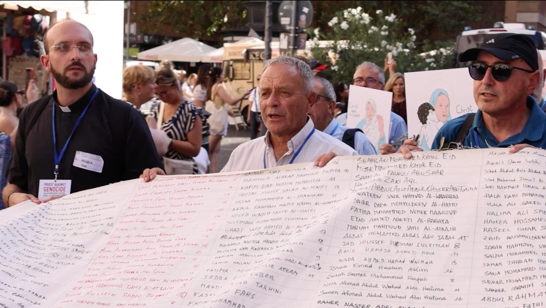 Three men hold a large sheet covered with handwritten names at a crowded public gathering.