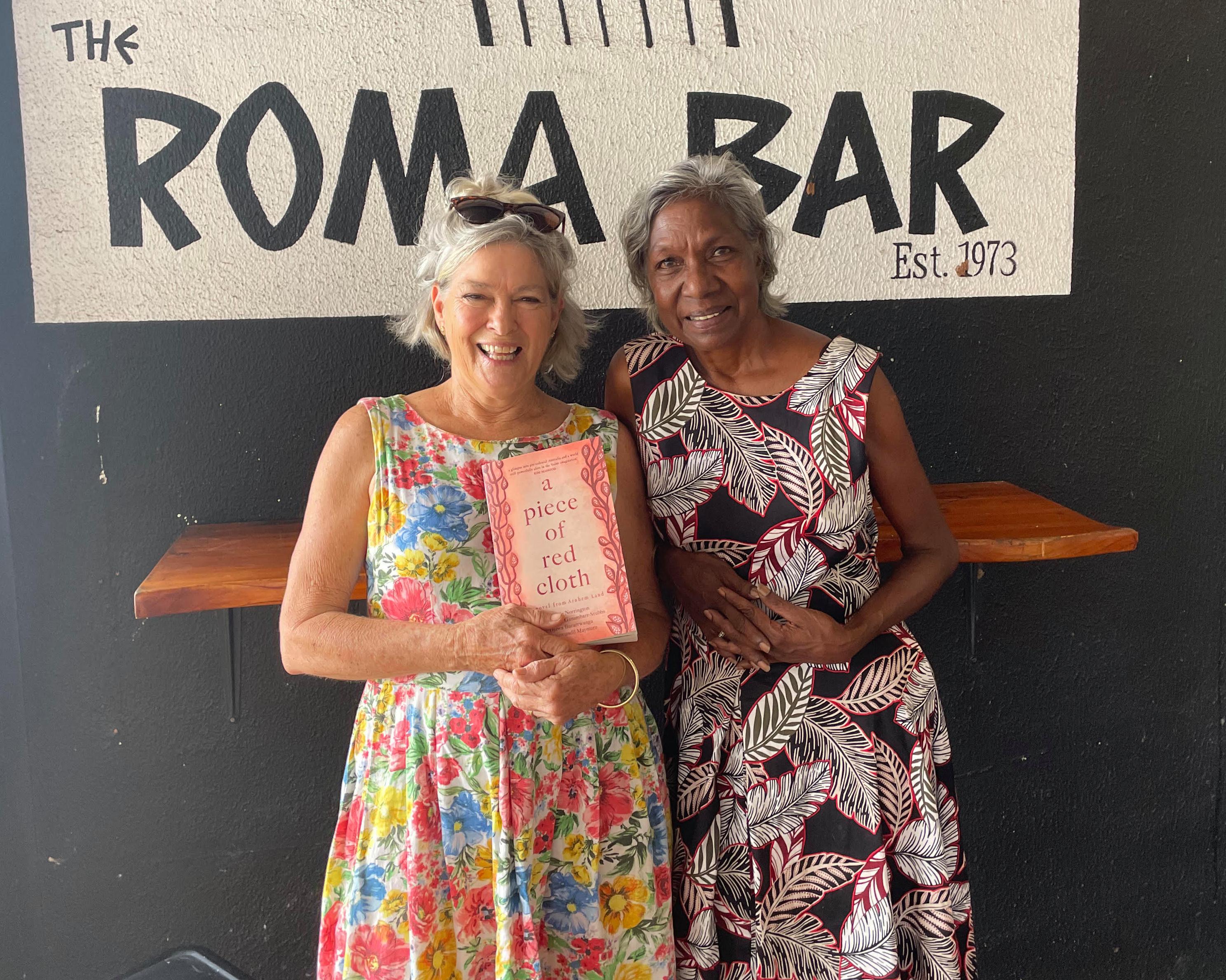 Two women stand smiling outside Roma Bar, one holding a copy of A Piece of Red Cloth.