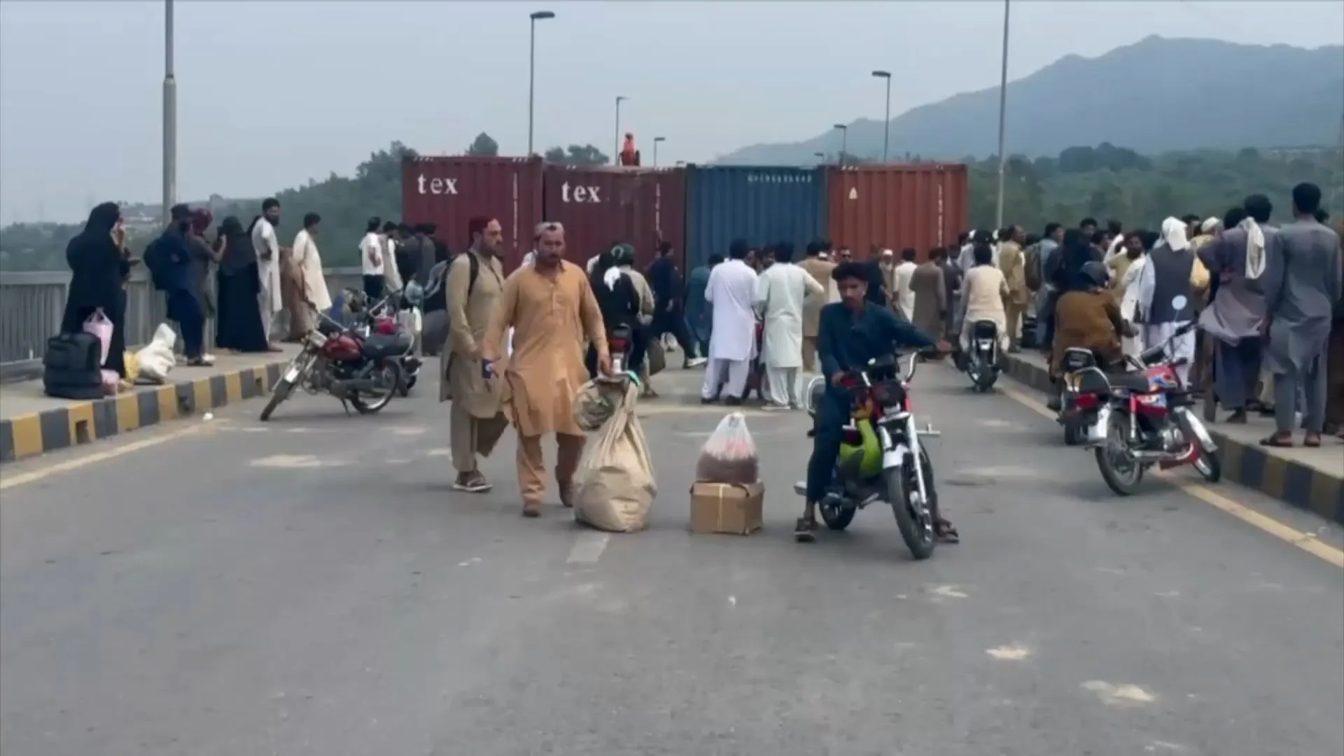 Crowd, motorcycles, and luggage gather on a bridge beside shipping containers with hills in the distance.
