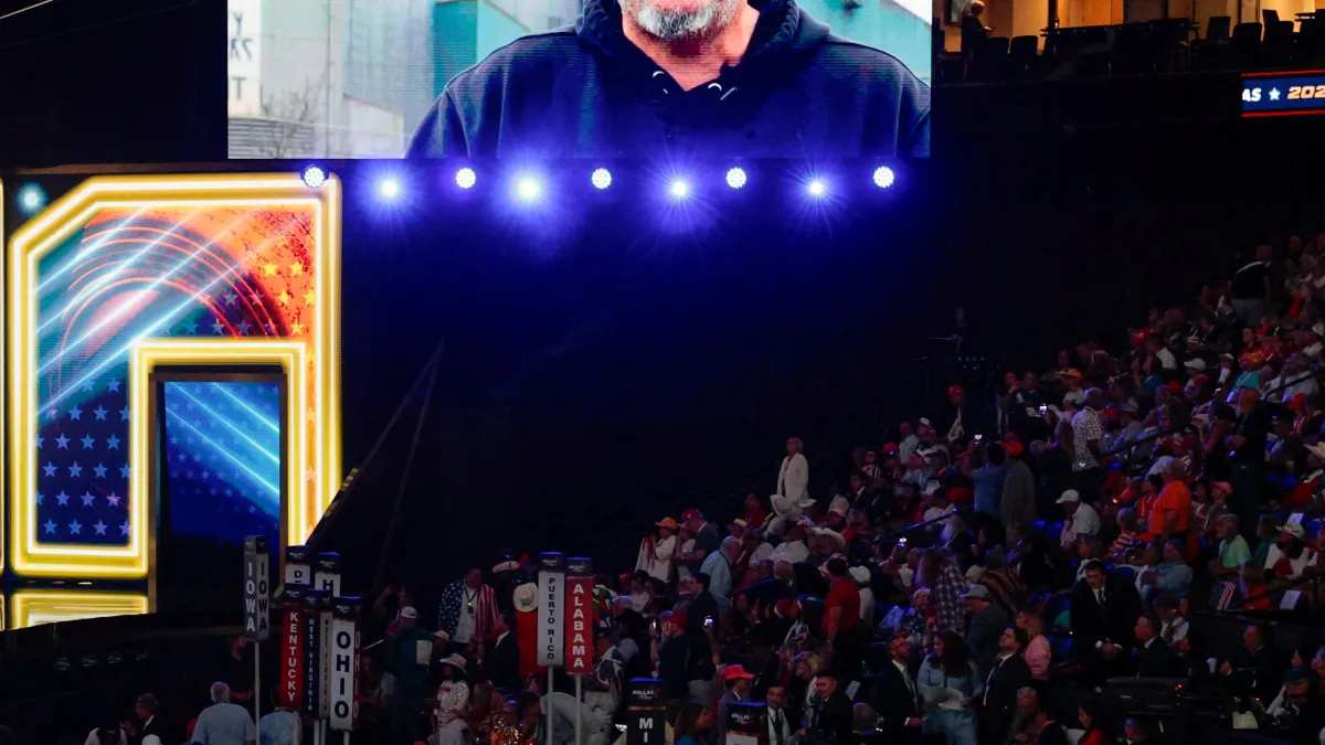 Convention audience watches a large video screen above a brightly lit stage with state delegation signs