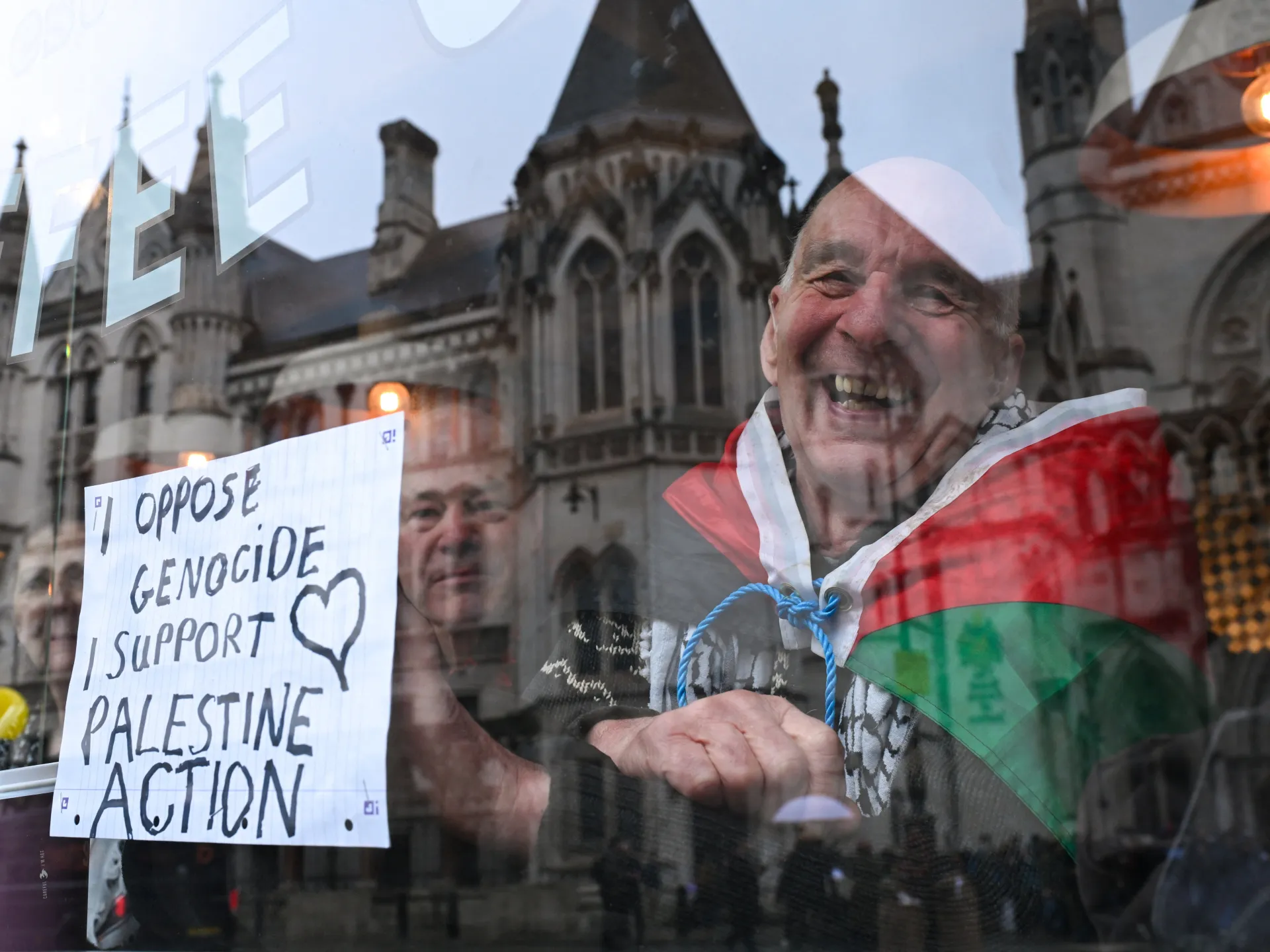 A smiling man in a Palestinian flag draped over his shoulders stands beside a handwritten protest sign.