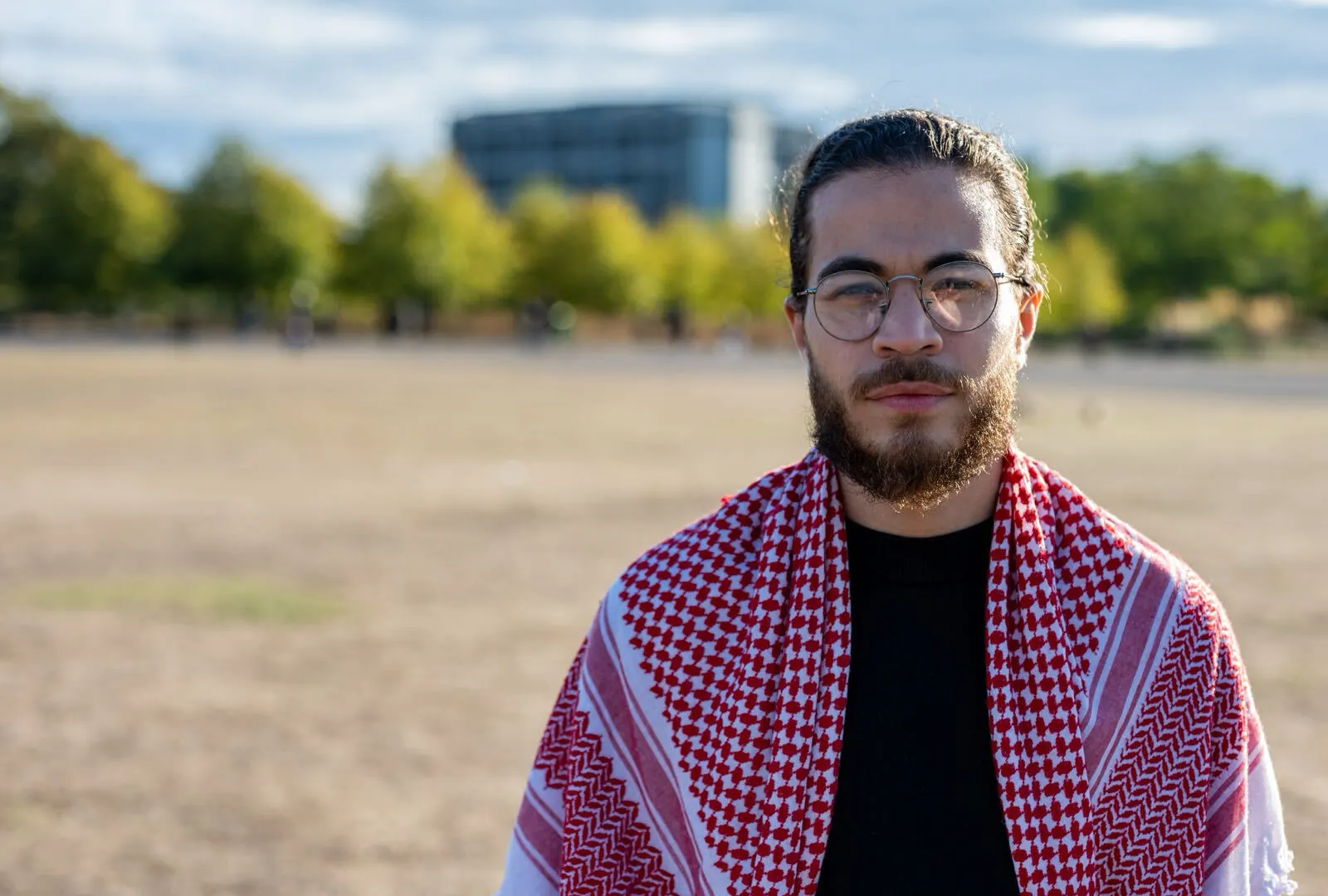 Bearded man wearing glasses and a red-and-white keffiyeh outdoors in a sunlit square