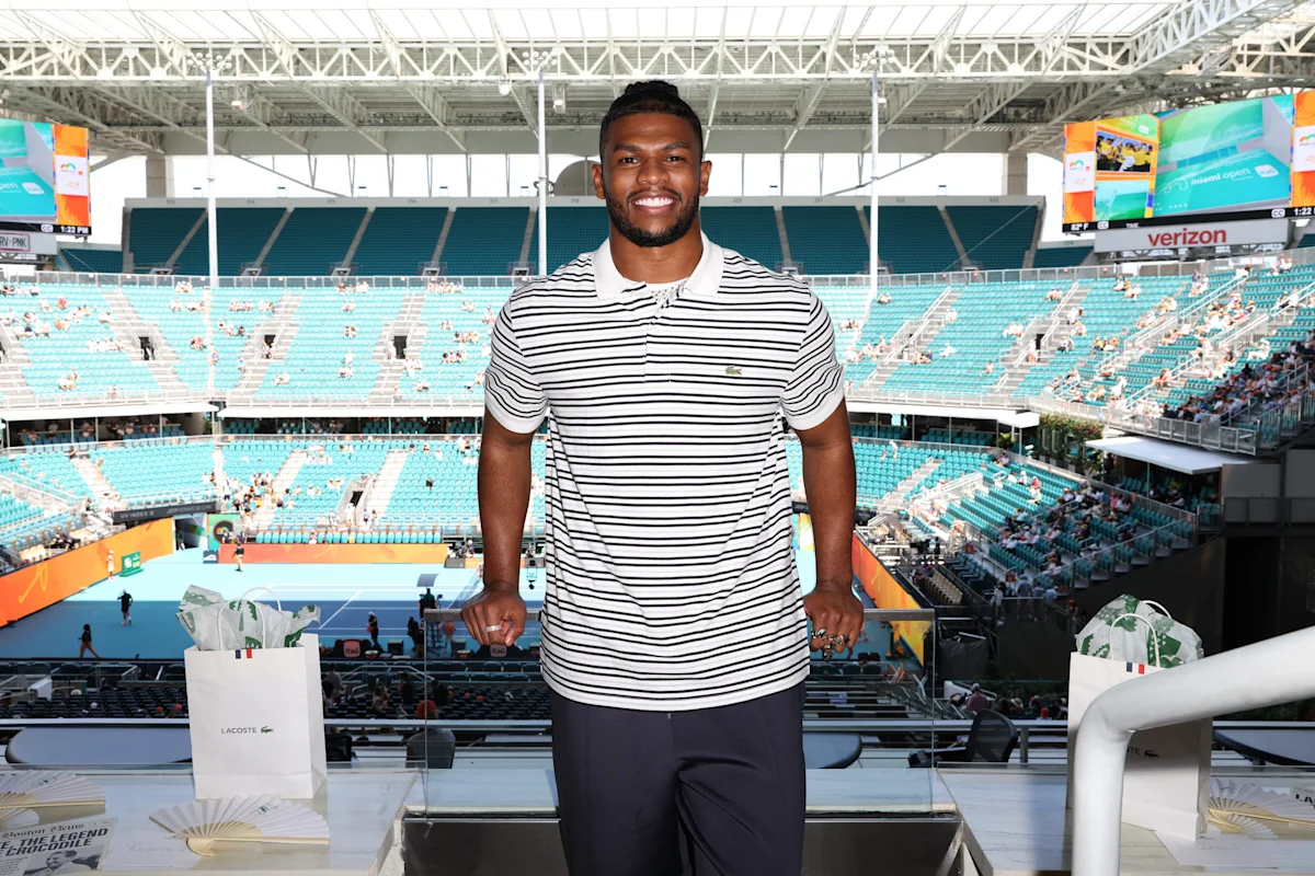 Smiling man in striped polo standing in a tennis stadium with turquoise seats