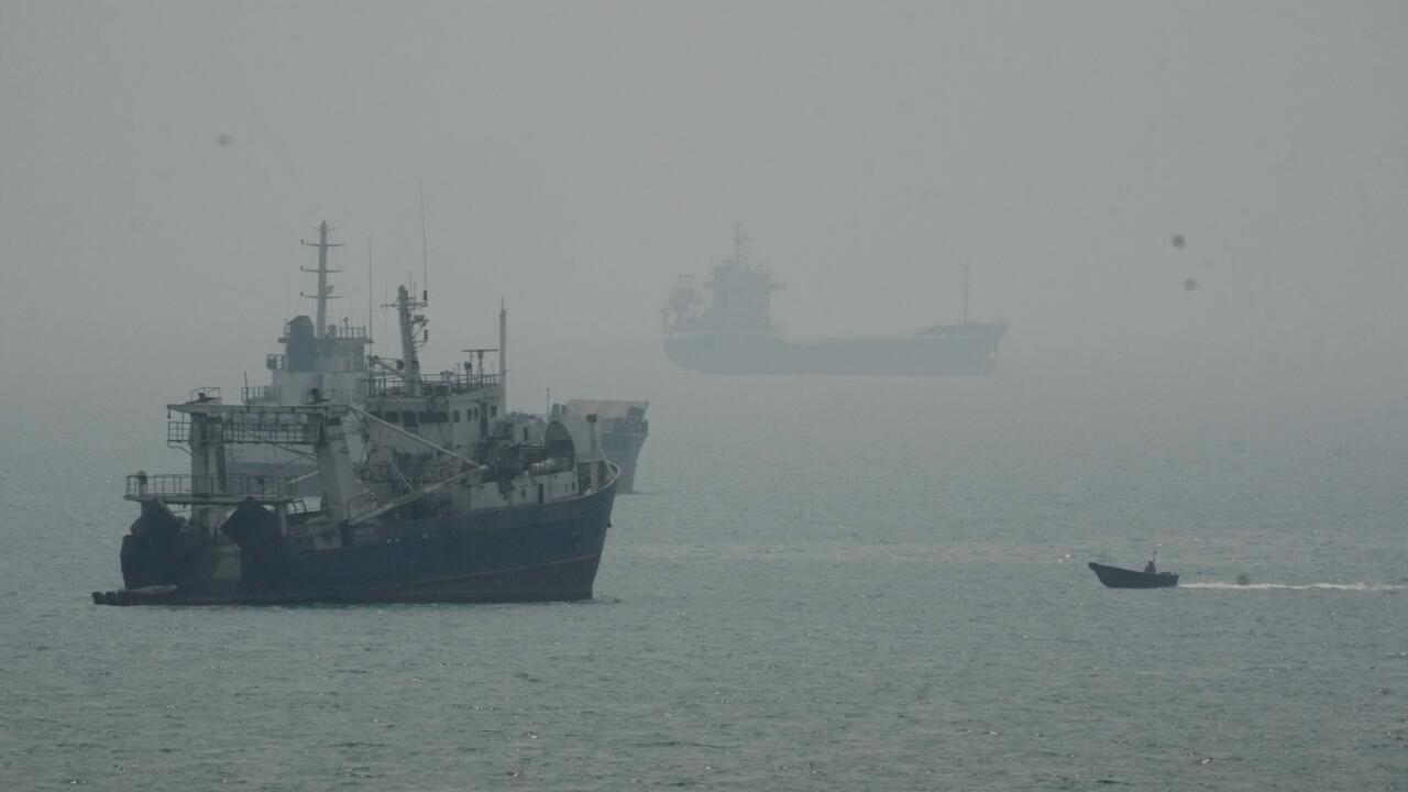 Fishing vessel in foreground, cargo ship shrouded in haze, and small boat crossing choppy water.