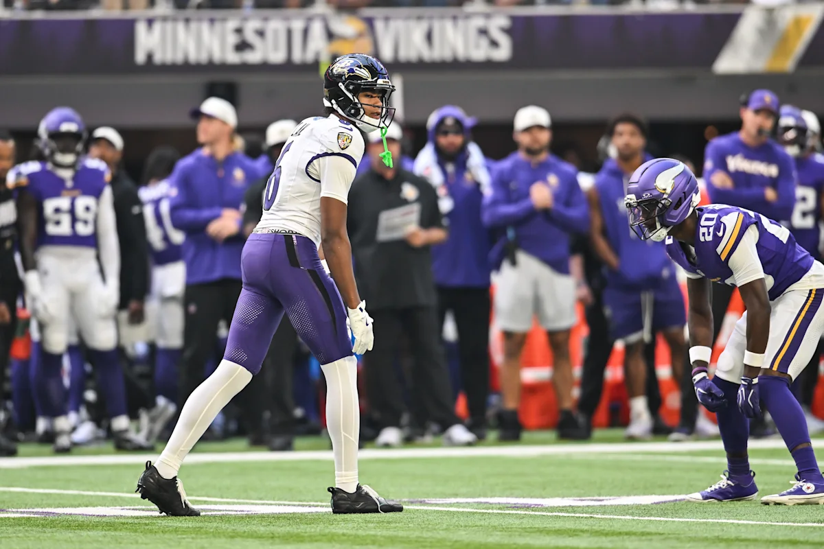 A football player in a white uniform faces two purple-clad defenders on the field.