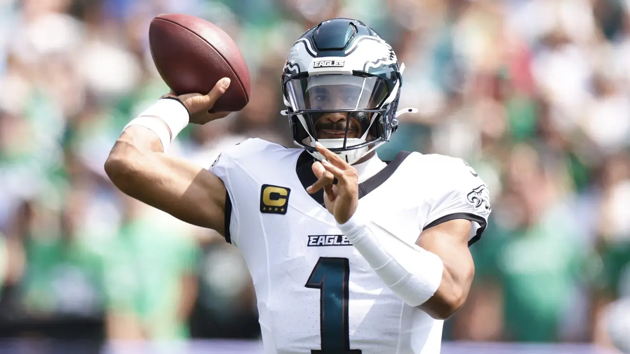 Eagles quarterback in a white uniform raises a football to throw against a blurred stadium crowd.