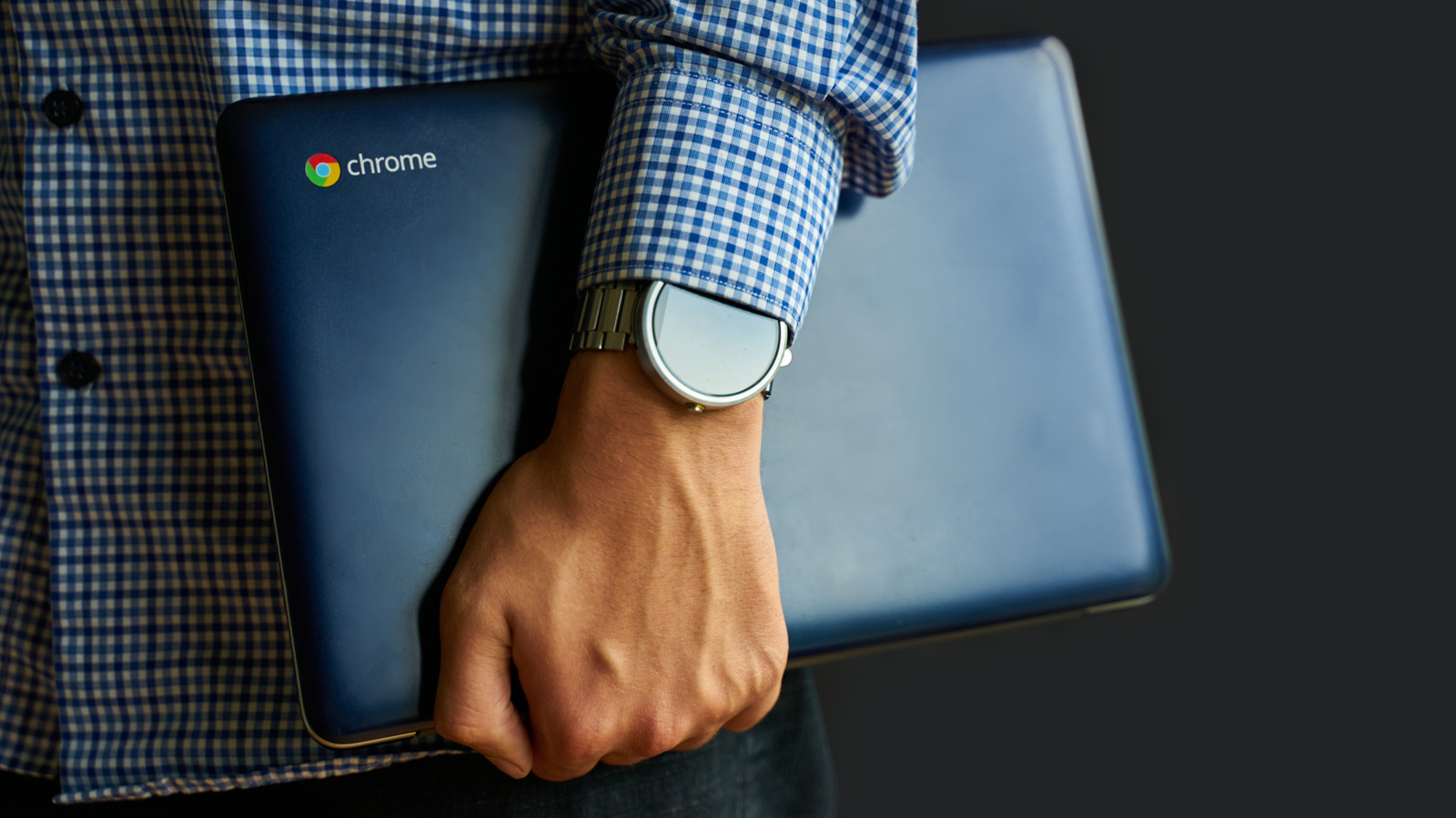 Person holding a closed Chromebook while wearing a checked shirt and wristwatch