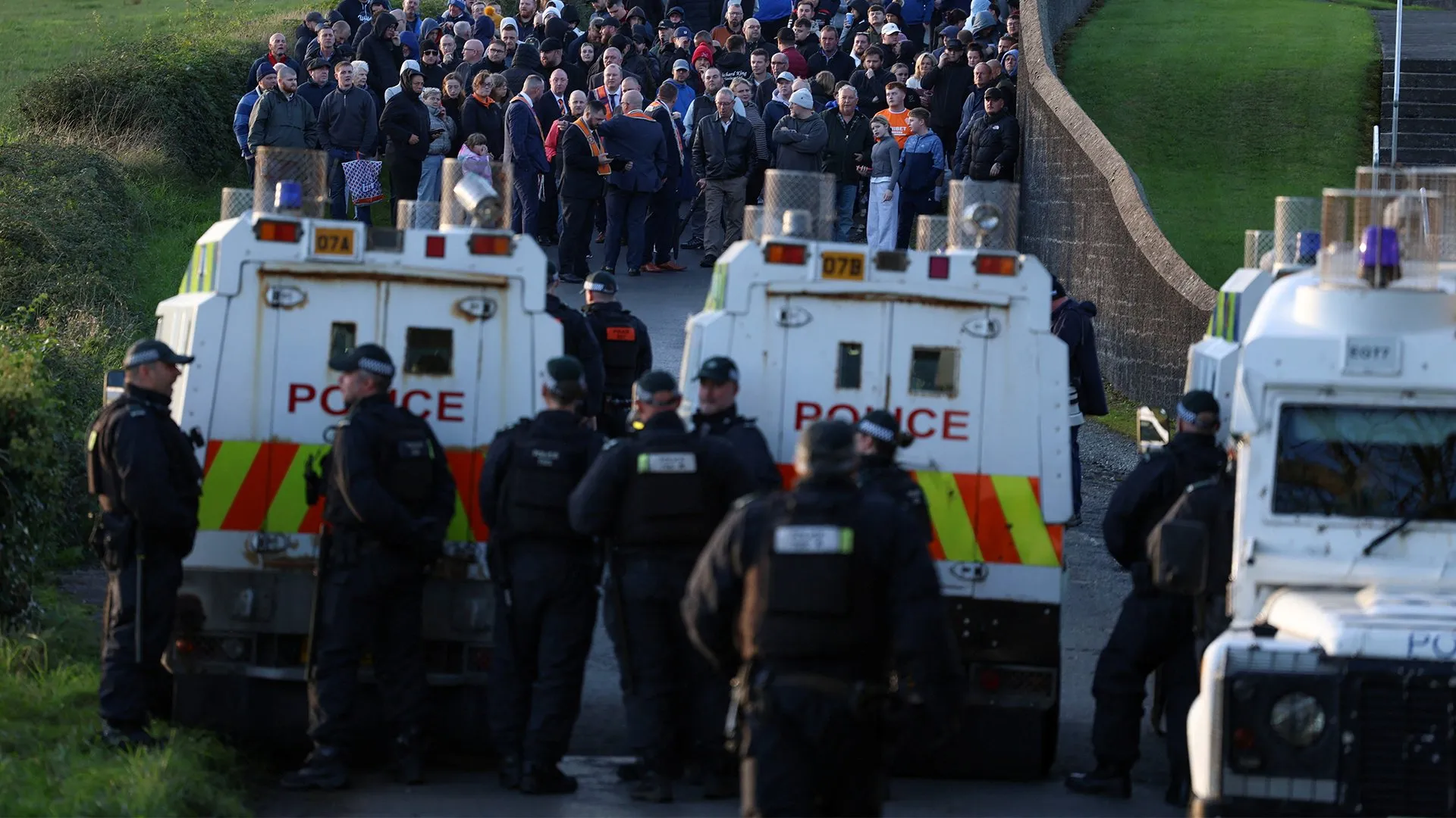 Police officers and vehicles face a large crowd gathered along a narrow road beside a grassy embankment.