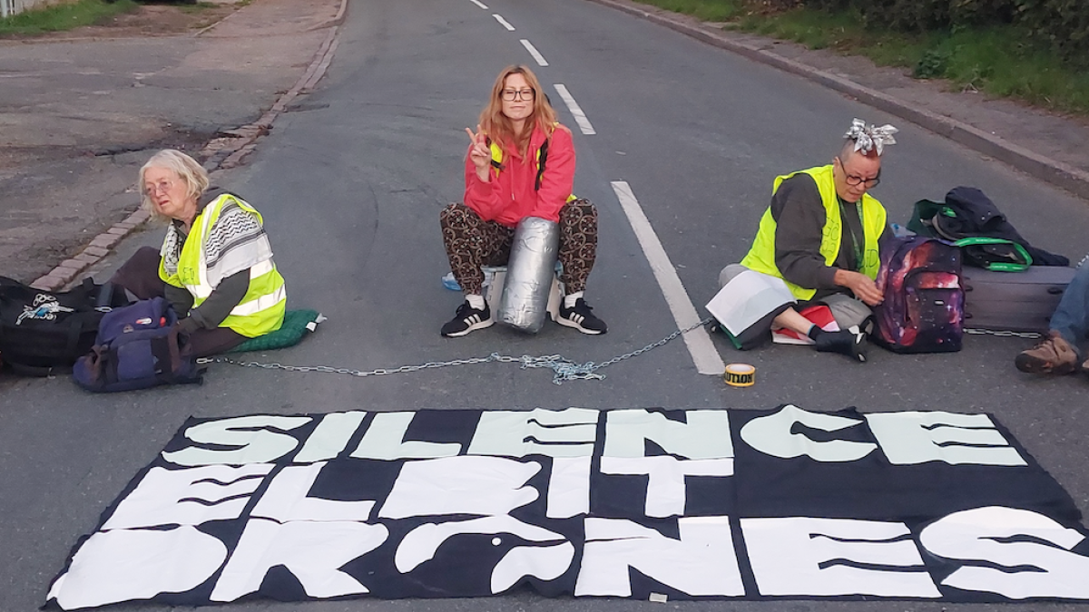 Three protesters sit across a road beside chains and a large black-and-white banner.