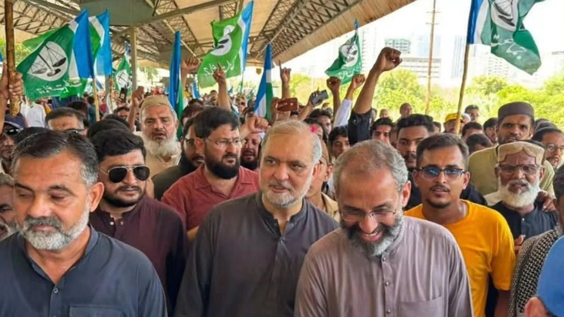 Crowd of men waving green and blue flags, with several raising fists during a protest.