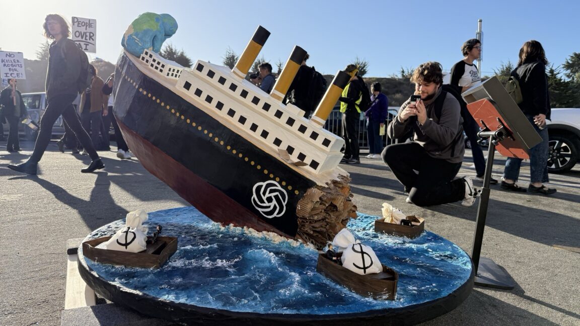 Protesters display a handmade model ship sinking beside small boats carrying money bags.